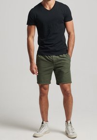 T-shirt nera a V, pantaloni corti verde oliva e sneaker bianche alte. L'outfit presenta una vestibilità casual con dettagli minimi.