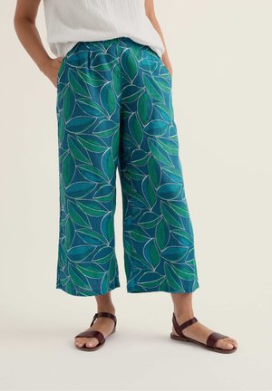 Vrouw draagt turquoise wijde broek met groene bladprint, wit mouwloos topje en bruine platte sandalen, staat met handen in de zakken.