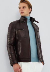 Giacca di pelle marrone con zip frontale, colletto alto, tasche laterali e dettagli trapuntati sulle spalle. Indossata sopra un dolcevita azzurro. 