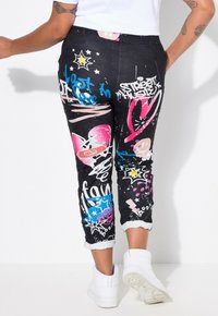 Pantalons en denim noir avec des motifs de graffiti colorés, incluant des cœurs, des étoiles et des textes. Les ourlets retroussés et une coupe ajustée complètent le look.