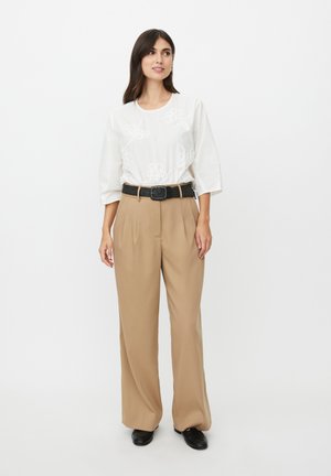 Mujer de pie llevando una blusa blanca bordada con flores, pantalones anchos beige con un cinturón negro y zapatos negros sobre un fondo liso.