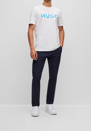 Homme portant un T-shirt blanc à manches courtes avec un logo bleu "HUGO", un pantalon bleu marine et des baskets blanches, debout avec une main dans la poche.