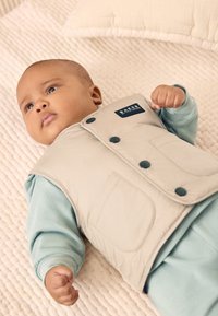 Veste matelassée beige avec des boutons foncés et un logo, portée par-dessus une chemise à manches longues bleu clair. Posée sur une surface texturée de couleur claire.