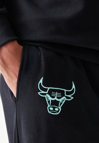 Μαύρο φόρμας παντελόνι με πλαϊνή τσέπη, διακοσμημένο με κεντημένο λογότυπο ταύρου σε ανοιχτό μπλε χρώμα. Το ύφασμα φαίνεται μαλακό και με υφή.