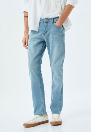 Jeans de mezclilla azul claro con un corte recto, diseño de cinco bolsillos y un ligero desgaste. Se llevan con una camisa blanca y zapatos informales.