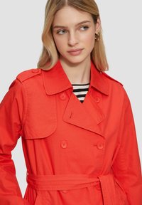 Sarkani tranšejera, izgatavota no gludas auduma, ar divām rindām pogām, regulējamu jostu un klasisku apkakles dizainu.