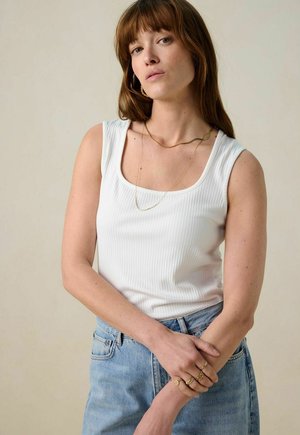White ribbed tank top with a square neckline, paired with light blue high-waisted jeans. Model wears gold layered necklaces and rings.