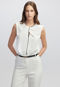 Mouwloze crèmekleurige blouse met geplooide details bij de hals, voorzien van een zwarte afwerking, gecombineerd met witte broeken en een zwarte riem met gouden beslag.