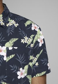 Camisa de manga corta en tela azul marino con un vibrante patrón floral que presenta flores blancas y amarillas, hojas verdes, y un bolsillo en el pecho.