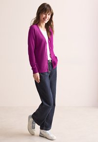 Gilet magenta avec fermeture à boutons, associé à un t-shirt blanc et un jean foncé, complété par des baskets bleu clair. Texture lisse, style décontracté.