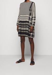 Schwarz-weiß gemustertes Kleid mit langen Ärmeln, das geometrische Designs und eine entspannte Passform aufweist, kombiniert mit schwarzen flachen Sandalen.