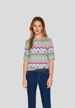 Bunt gemustertes Oberteil mit Zickzackmuster in Grüntönen, Lila und Blau; kurze Ärmel; Rundhalsausschnitt; kombiniert mit dunkelblauen Hosen.