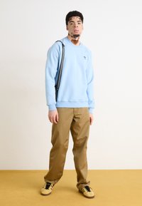 Svetlo modra dukserica z ribastimi manšetami in obodom, v kombinaciji z bež hlačami in rumenimi športnimi copati. Model drži črno torbico na naramnice.