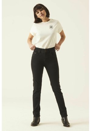 T-shirt en coton blanc avec un logo noir sur la poitrine, associé à un jean noir taille haute. Modèle portant des bottines noires.