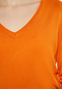 Narancssárga V nyakú pulóver bordázott textúrával és enyhén slim szabású ujjal. Az anyag lágy és könnyűnek tűnik. Közelkép a nyakrészről.
