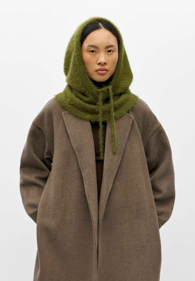 Femme portant un manteau marron oversize et une écharpe à capuche verte et duveteuse nouée sous le menton, debout devant un fond clair uni.