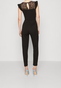 Svart jumpsuit med spetsdetaljer på baksidan, korta volangärmar och figurnära ben. Matchad med ljusblå högklackade skor.