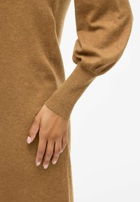 Brown knitted dress featuring a ribbed cuff and a voluminous sleeve. Soft texture, fitted at the wrist, with a smooth body.