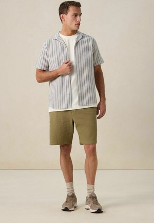 Chemise à manches courtes à boutons avec des rayures bleues et blanches sur un t-shirt blanc, associée à un short en olive texturé et des baskets beiges.