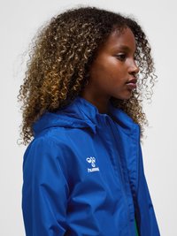 Ung person med krøllet hår iført en blå Hummel lynlåsjakke, set mod højre på en ensfarvet lys baggrund.