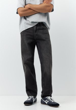 Mann steht mit verschränkten Armen, trägt ausgewaschene schwarze Jeans, graues T-Shirt und schwarz-weiße Turnschuhe vor einfachem Hintergrund.