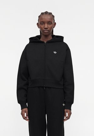 Osoba s vrkočmi, ktorá má na sebe čiernu mikinu na zips a čierne nohavice s malými bielymi logami Adidas na hrudi a stehne.