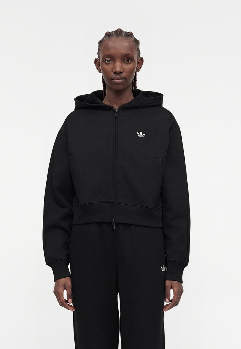Personne aux cheveux tressés portant un sweat à capuche noir zippé et un pantalon noir assorti avec petits logos blancs Adidas sur la poitrine et la cuisse.