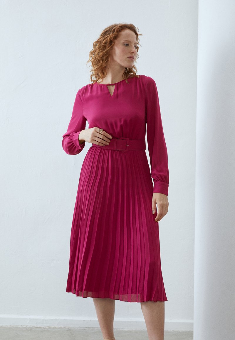 Rochie midi pliată dintr-un material roz aprins, cu mâneci lungi, decolteu tip eyelet și o curea lată la talie pentru definiție.