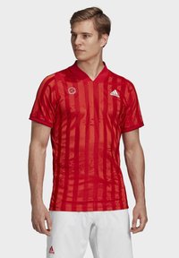 Jersey atlético rojo con rayas verticales, mangas cortas y cuello en forma de V. Presenta el logo de Adidas y un emblema redondo en el lado izquierdo del pecho.
