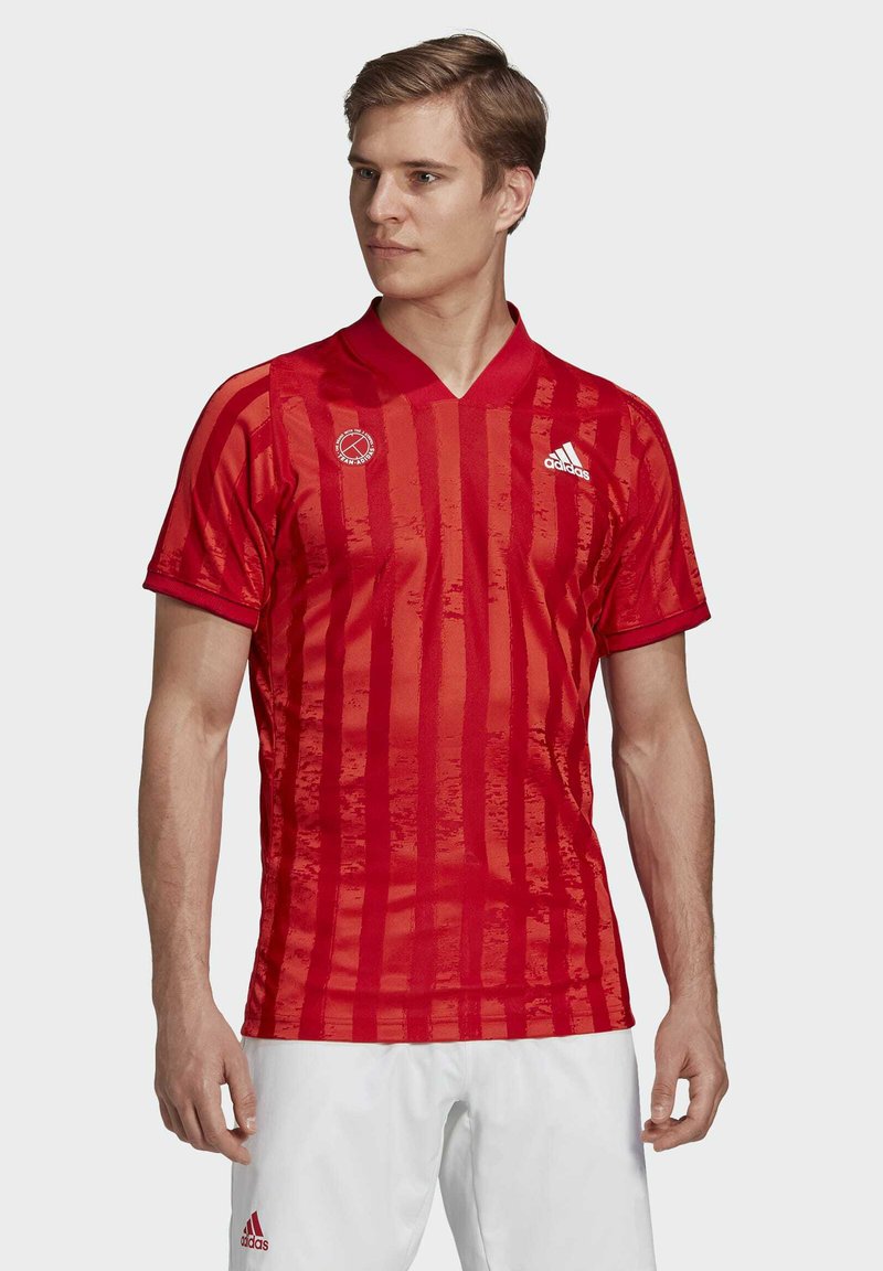 Jersey atlético rojo con rayas verticales, mangas cortas y cuello en forma de V. Presenta el logo de Adidas y un emblema redondo en el lado izquierdo del pecho.