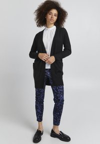 Črna cardigan jakna z odprtim sprednjim delom in žepi, nošena čez belo bluzo z visokim ovratnikom. V kombinaciji z modrimi hlačami z leopardjim vzorcem in črnimi loaferji.