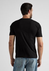 T-shirt en coton noir à manches courtes, avec un col rond et une texture lisse, vu de dos. Porte un jean en denim bleu clair.