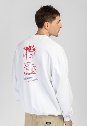 Man wearing white sweatshirt with red graphic of smiling coffee cup and text "be a barista!" on back, beige pants, looking sideways.