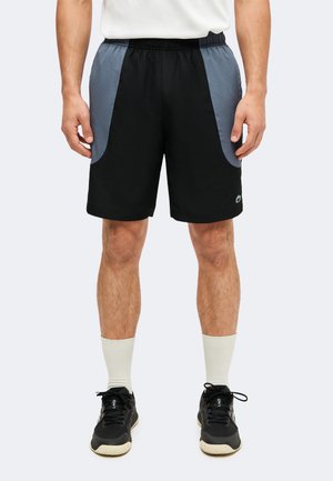 Mann trägt schwarze Shorts mit grauen Seitenpaneelen, weiße Socken und schwarze Sportschuhe und steht vor einem schlichten Hintergrund.
