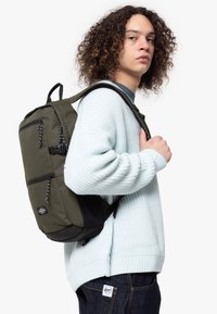 Sac à dos en tissu vert avec plusieurs poches extérieures, accents noirs et bretelles à motifs, porté par-dessus un pull en tricot bleu clair.