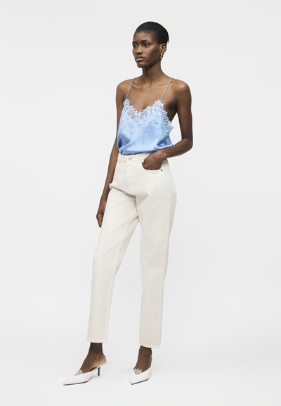 Ženska, oblečena v modro satenasto majico z čipkastimi detajli, bele hlače z visokim pasom in bele konične pete, stoji z eno roko v žepu.