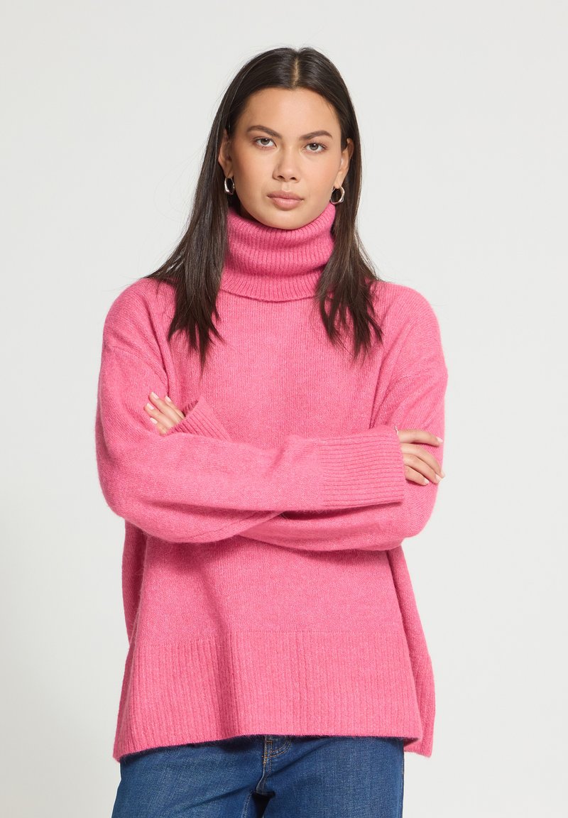 Jeune femme aux longs cheveux foncés, portant des boucles d'oreilles créoles, un pull à col roulé rose et un jean bleu, debout les bras croisés.