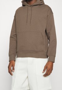 Brown hooded sweatshirt with a front pocket, made from a textured fabric, featuring adjustable drawstrings and a relaxed fit. White shorts.