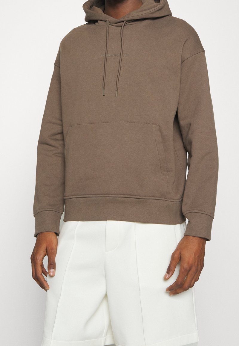 Brown hooded sweatshirt with a front pocket, made from a textured fabric, featuring adjustable drawstrings and a relaxed fit. White shorts.
