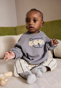 Felpa grigia con design di orso, pantaloni a righe crema e calzini grigi a fantasia. Bambino seduto su una superficie chiara, giocattolo di legno nelle vicinanze.