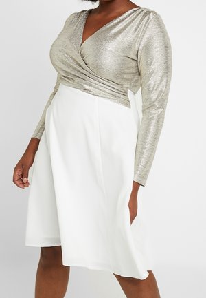Frau trägt ein silbernes, metallisches Wickeloberteil mit langen Ärmeln, kombiniert mit einem hoch taillierten weißen Rock, steht vor einem schlichten Hintergrund.