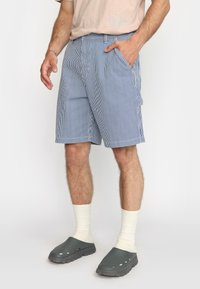 Heren blauwe en witte gestreepte shorts met zijzakken, gemaakt van lichtgewicht materiaal. Gecombineerd met grijze klompen en lichte geribbelde sokken.