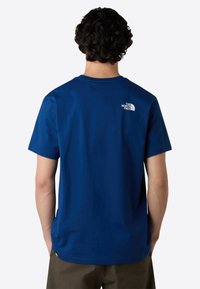 Námořnická modrá bavlněná tričko s krátkými rukávy, kulatým výstřihem a bílým logem vytištěným na horní části zad. Hladká textura látky.