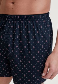 Marineblauwe boxers gemaakt van zachte stof, met een patroon van kleine diamanten en ovalen in lichtroze en wit. Elastische tailleband met knopen.