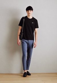 Svart T-shirt med kort ärm, ljusblå skräddarsydda byxor och svarta skor. Modellen håller en svart axelväska och står framför en neutral bakgrund.