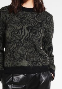 Czarny i zielony sweter w wzory z fakturowaną powierzchnią. Posiada okrągły dekolt oraz ściągacze przy mankietach i dolnym brzegu. Połączony z czarnymi skórzanymi spodniami.