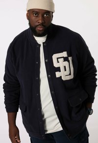Navy Fleecejacke im Baseball-Stil mit Druckknöpfen und großem weißen "SU"-Logo auf der Brust. Lange Ärmel und Seitentaschen.
