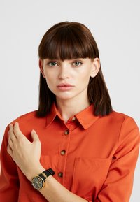 Fekete óra arany kiegészítőkkel, fekete szíjon, egy kézen viselve. A téma narancssárga gombos inget visel, két mellzsebbel.