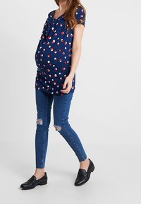Top de maternidad en azul marino con puntos rojos y blancos, de manga corta y escote en V, combinado con jeans ajustados azules y mocasines negros.