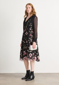 Schwarzes Blumenkleid mit transparenten Ärmeln, verziert mit rosa und grünen Mustern. Kombiniert mit einer silbernen Clutch und schwarzen Stiefeletten.